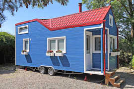 Ein kleines blaues Wohnhaus mit weißen Fensterläden, rotem Dach und Rädern steht auf einer Kiesfläche