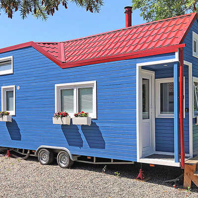Ein kleines blaues Wohnhaus mit weißen Fensterläden, rotem Dach und Rädern steht auf einer Kiesfläche