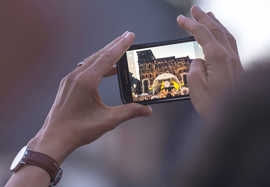 Zwei Hände halten ein Smartphone,.auf dessen Display die Porta Nigra mit Konzertbühne zu sehen ist.
