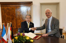 Wolfram Leibe und Tadahiro Matsubara sitzen an einem mit Blumen und Fahnen geschmückten Tisch und reichen sich die Hände.