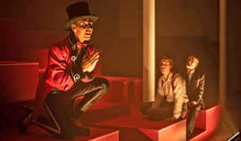 Szene einer Theateraufführung: Im Vordergrund kniet ein Schauspieler mit schwarzem Zylinder, rotem Sakko und schwarzer Hose, im Hintergrund sitzen zwei weitere Darsteller auf roten Stufen.