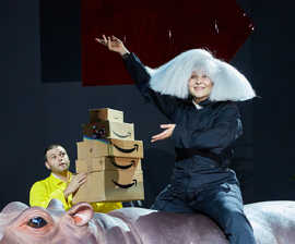 Die groteske Szene einer Theateraufführung zeigt eine Frau mit ausladender weißer Perücke, die auf einer Nilpferdattrappe reitet und einen Mann, der einen Stapel Amazon-Pakete trägt.