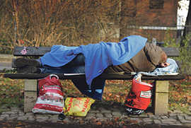 Eine Person liegt zugedeckt auf einer Parkbank, vor ihr auf dem Boden volle Plastiktüten