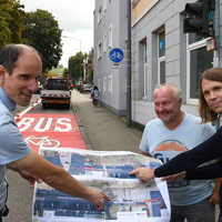 Dre Personen halten einen Papierbogen mit Planzeichnungen, im Hintergrund ist ein rot eingefärbter Straßenabschnitt mit dem Schriftzug Bus und einem Fahrradsymbol.