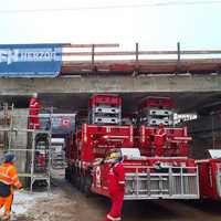Der Überbau einer Brücke wird auf einer Baustelle mit Arbeitern und Ingenieuren in roten und orangefarbenen Overalls mit Hilfe von Spezialfahrzeugen an ihren Platz gehoben. 