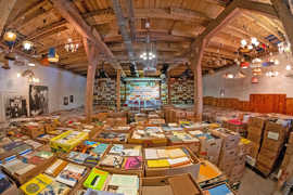 In einem saal mit Balkendecke udn verschiedenartigen Deckenlampen stehen mit Büchern gefüllte Kisten und Kartons 
