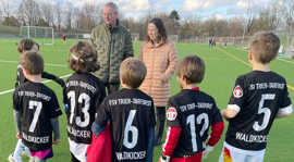 Zwei Erwachsene sprechen auf einem Kunstrasenplatz mit fünf Kindern, die Fußballtrikots tragen