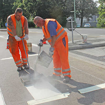 Mitarbeiter einer Baufirma aus Neunkirchen gießen die Kunststoffmasse für die Zebrastreifen auf dem erneuerten Übergang an der Kreuzung Hans-Böckler-Allee/Tessenowstraße. 