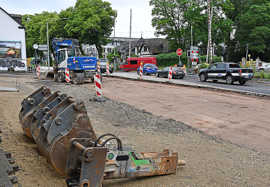 Blick auf eine Straßenbaustelle mit einem Bagger und weiteren Gerätschaften. Der Verkehr fließt zweispurig an der Baustelle vorbei.