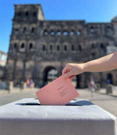 Vor dem Hintergrund der Porta Nigra wird ein Wahlbrief in eine Urne gesteckt.