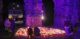 Menschen knien vor der Porta Nigra und entzünden Kerzen. Das Wahrzeichen der Stadt wird in der Dunkelheit aktuell mit der Trauerfarbe violett angestrahlt.