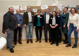 Gruppenfoto von neun Personen vor einer Plakatwand.
