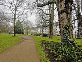 Ein Weg führt durch einen Park mit Wiesen und Bäumen, im hintergrund ist dichte Wohnbebauung zu sehen.