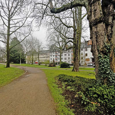 Ein Weg führt durch einen Park mit Wiesen und Bäumen, im hintergrund ist dichte Wohnbebauung zu sehen.