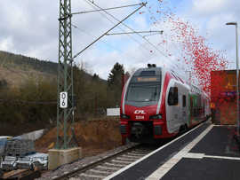 Ein rot-weißer Doppelstockzug fährt an einem Bahnsteig entlang und wird mit einer rote Konfettiwolke beschossen