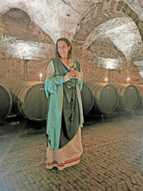 Ein Frau trägt ein langes Kleid in Grüntönen und steht mit einem Weinglas in der Hand vor Weinfässern in einem Kellergewölbe.