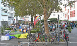 Auf einer asphaltierten Freifläche stehen Bäume, Fahrräder, Sitzkissen, Sitzbänke, eine Fahne und Tische, an denen sich Besucher unterhalten