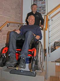 Andreas Rump wird von Jürgen Wingertszahn in einem Rollstuhl mit dem darunter montierten Steiger in den ersten Stock befördert.