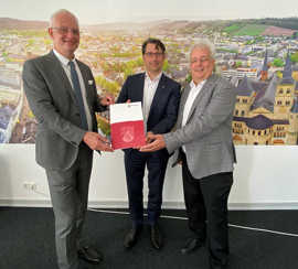 OB Wolfram Leibe, Staatssekretär Michael Hauer und Baudezernent Andreas Ludwig stehen vor einem großen Foto von Triers Innenstadt und präsentieren den Förderbescheid.