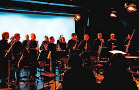 Ein schwarz gekleidetes Ensemble mit verschiedenen Blasinstrumenten steht auf der Bühne