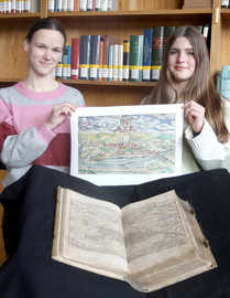 Zwei junge Frauen präsentieren eine historische Stadtansicht von Trier. Vor ihnen liegt aufgeschlagen eine Ausgabe der Cosmographia