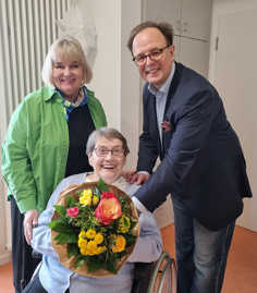 Anna Cäcilia-Klasen im Rollstuhl lacht und hält einen Blumenstrauß in den Händen, hinter ihr stehen Petra Block und Ralf Britten