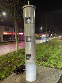 Eine Blitzersäule steht neben einer Straße in einem Grünstreifen, abgeschlagene Glasteile liegen daneben.