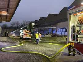 Man sieht eine Lagerhalle rechts im BIld. Hinten ist ein Dachstuhl, auf dem Feuer zu sehen ist. Davor sind zwei Feuerwehrfahrzeuge. Feuerwehrmänner sind mit Löscharbeiten beschäftigt.