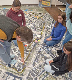 Auf einer mit einem Luftbild von Trier bedruckten Lkw-Plane platzieren Teilnehmerinnen eines Workshops verschiedene Markierungen