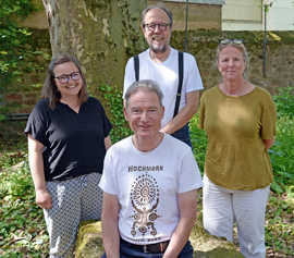 Das Gruppenbild zeigt zwei Männer (davon einer sitzend) und zwei Frauen in sommerlicher Kleidung in einem Park.