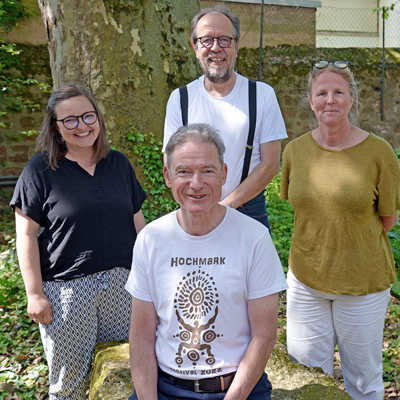 Das Gruppenbild zeigt zwei Männer (davon einer sitzend) und zwei Frauen in sommerlicher Kleidung in einem Park.