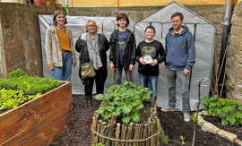 Zwei Jugendliche und drei Erwachsene stehen in einem Garten mit verschiedenen Gemüsebeeten und präserntieren das Gartenpatenschild.