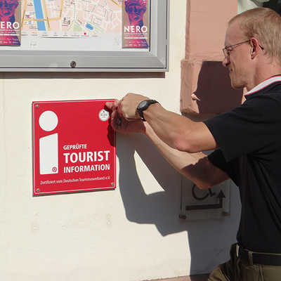 Alexander Geißler montiert das neue rote Schild mit der i-Marke in der Nähe des Eingangs zur Trierer Tourist-Info neben der Porta Nigra. Foto: ttm