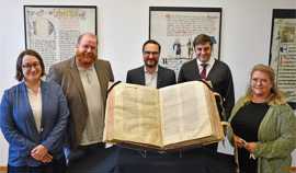 Alt-Text: Magdalena Palica, Leiterin der Wissenschaftlichen Bibliothek Trier (rechts), und Kulturdezernent Markus Nöhl (2. von rechts) versammeln sich mit Dr. Claude D. Conter, Direktor der Luxemburgischen Nationalbibliothek (Mitte), PD Dr. Francesco Roberg, Direktor der Wissenschaftlichen Bibliothek Trier (2.v.r.), sowie der Digitalisierungsbeauftragten Anja Runkel (ganz rechts) um die aufgeschlagene Riesenbibel.