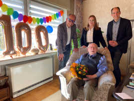Herbert Schwarz, der 100 Jahre alt geworden ist, sitzt in einem Sessel in seinem Wohnzimmer. Links neben ihm am Fenster hängen drei Luftballons, die eine 100 zeigen. Hinter ihm stehen Olewigs Ortsvorsteher Dr. Lorenz Fischer, Enkeltochter Hannah und Dezernent Dr. Thilo Becker.