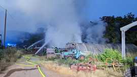 Das Bild zeigt im Vordergrund landwirtschaftliche Maschinen und einen Pflanzentunnel. Im Hintergrund löschen die Feuerwehrleute den qualmenden Holzstapel.