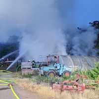 Das Bild zeigt im Vordergrund landwirtschaftliche Maschinen und einen Pflanzentunnel. Im Hintergrund löschen die Feuerwehrleute den qualmenden Holzstapel.
