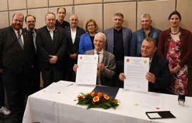 Wolfram Leibe und Valerii Marchenko sitzen an einem blumengeschmückten Tisch und präsentieren zwei Urkunden. Hinter ihnen stehen weitere Repräsentantinnen und Repräsentanten.