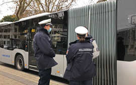 Zwei Kontrollkräfte des Kommunalen Vollzugsdienstes kontrollieren die Nachweise eines Fahrgastes. Im Hintergrund steht ein Bus.