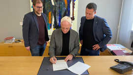 Wolfram Leibe sitzt an seinem Schreibtisch und unterschreibt einen Vertrag. Zwei Mitarbeiter stehen rechts und links hinter ihm.