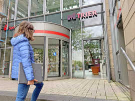 Eine Frau trägt einen Aktenordner und geht auf eine Eingangstür in einer Gasfront, an der das Logo der Stadt Trier angebracht ist