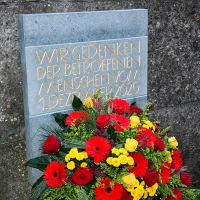 Die Inschrift der Gedenktafel aus Kalksandstein lautet: "Wir gedenken der betroffenen Menschen vom 1. Dezember 2020". Die Schrift ist golden. Davor steht ein großer Blumenstrauß mit roten und gelben Blumen.