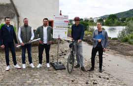 Fünf Projektbeteiligte stehen an einem gepflasterten Abschnitt des Radwegs am Moselufer und präsentieren eine Tafel mit Informationen über Sperrungen und Umleitungen.