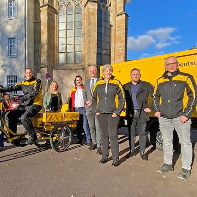 Die Deutsche Post DHL hat unter anderem E-Trikes und Elektro-Lastwagen im Einsatz. Vor dem Trierer Rathaus präsentieren Tanja Lauer (ver.di), Julia Hollweg (Klimaschutzmanagerin Stadt Trier), Zustellerin Katja Rößel, MdB Verena Hubertz, Thomas Schneider (Post-Betriebschef), OB Wolfram Leibe, Anke Podewin, (Niederlassungsleiterin Koblenz), Sylvester Swierczynski (Vorsitzender Betriebsrat), Matthias Petersmarck (Zustellungsleiter für Trier) und Dirk Kessler (Niederlassungsleiter Multikanalvertrieb) die umweltfreundlichen Fahrzeuge.