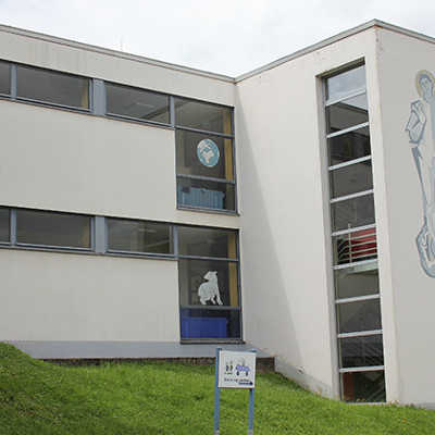 Das Irscher Gebäude wurde im Herbst 1962 als katholische Volksschule eingeweiht und erhielt den Namen des heiligen Georg, der rechts auf der Fassade zu sehen ist. 
