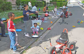 Auf einer Straße sind viele Kinder und unterschiedliches Spielzeug zu sehen. Zwei Kinder stehen im Vordergrund und versuchen, mit Stöcken und Schlingen riesige Seifenblasen zu machen.