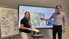 Thilo Becker und Julia Hollweg stehen vor einem großen Bildschirm, auf dem ein Ausschnitt eines Stadtplans zu sehen ist. 
