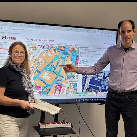 Thilo Becker und Julia Hollweg stehen vor einem großen Bildschirm, auf dem ein Ausschnitt eines Stadtplans zu sehen ist. 
