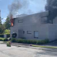 Das Bild zeigt ein zweigeschossiges Haus mit Flachdach. Links schlagen Flammen aus dem Haus, rechts dichter schwarzer Rauch.