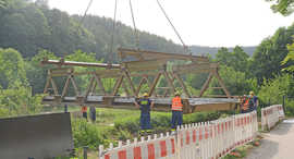 Fachleute des THW nehmen die vormontierte Brücke, die mit einem Kran von der Straße ans Flussufer gehievt wird, „in Empfang“.